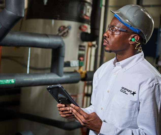 Worker in safety gear and an Aramark uniform examining pipes and holding a tablet.